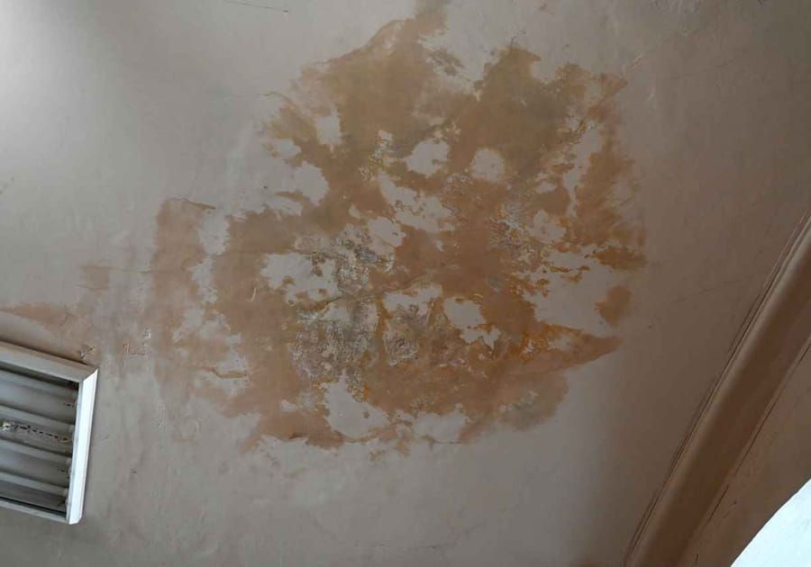 Stains and peeling paint are visible on a ceiling, indicating water damage. The area is well-lit by natural daylight, highlighting the uneven texture